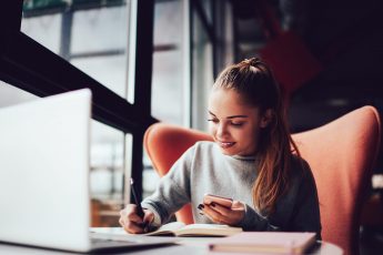 Smiling female student copying information from received message getting discount code for online shopping.Prosperous business woman satisfied with calculation of income sitting in cafe interior